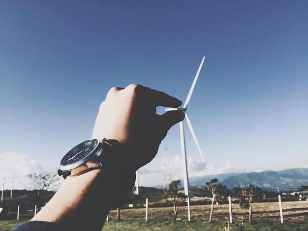 A detailed close-up of a hand holding a small solar panel, with a blurred background showing a wind turbine farm. The image is symbolic of individual and corporate investment in renewable energy.