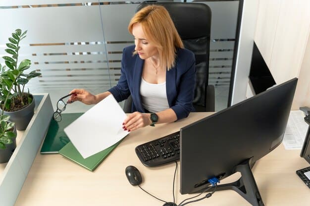 A person sitting at a desk, reviewing documents related to benefits program requirements, with a computer screen showing a checklist of tasks.