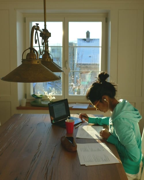 An international student looking concerned while reviewing bills and financial documents at a table in a dorm room.